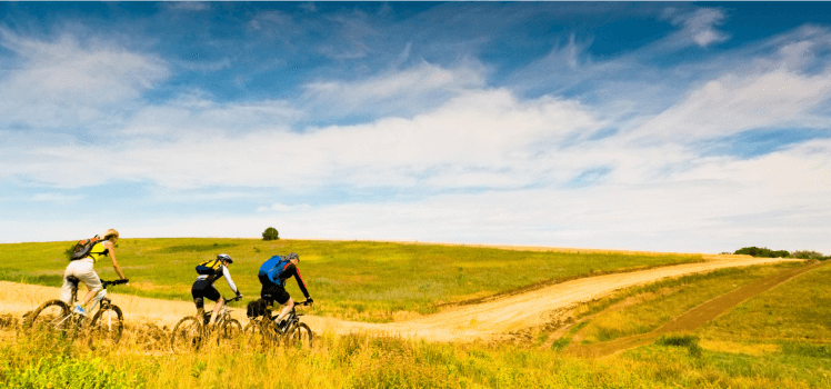 Three people biking on a dirt trail through a field under a cloudy blue sky.