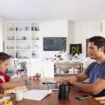 Man in blue sitting at a home dining table working remotely from a laptop while his son colors across the table.