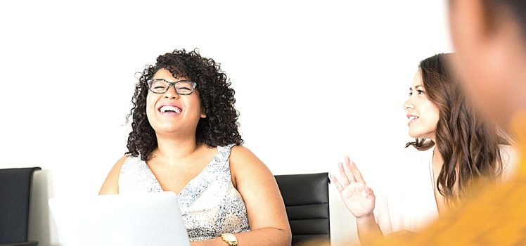 Woman with curly hair and glasses with a laptop laughing.