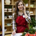 Woman in a plain shirt and red apron on the phone taking an order in a flower shop.