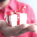 Closeup of a person in a pink shirt holding a very small white box with red ribbon and white hearts.