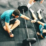 Overhead view of two men doing a fist bump over a large barbell as they sit or lay on the ground.