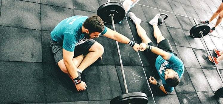 Overhead view of two men doing a fist bump over a large barbell as they sit or lay on the ground.