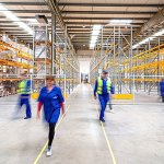 Multiple warehouse workers in blue with yellow caution vests walking through an empty warehouse. wellness programs improve employee retention