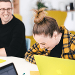 Two people sitting at a desk laughing and working on laptops.