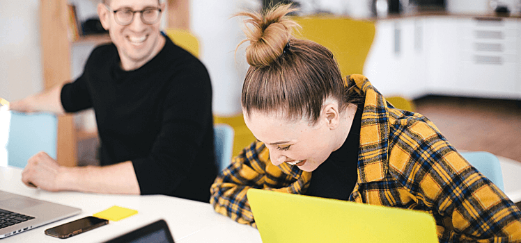 Two people sitting at a desk laughing and working on laptops.