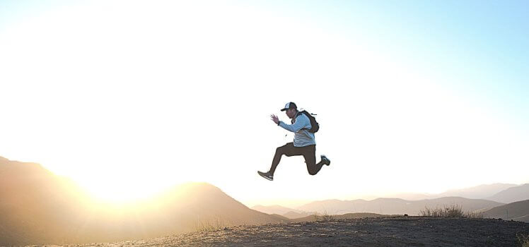 Man in a hat on a trail jumping high into the air as if he is floating in front of a large mountain range. 10 Effective Ways to Motivate Employees in the Workplace