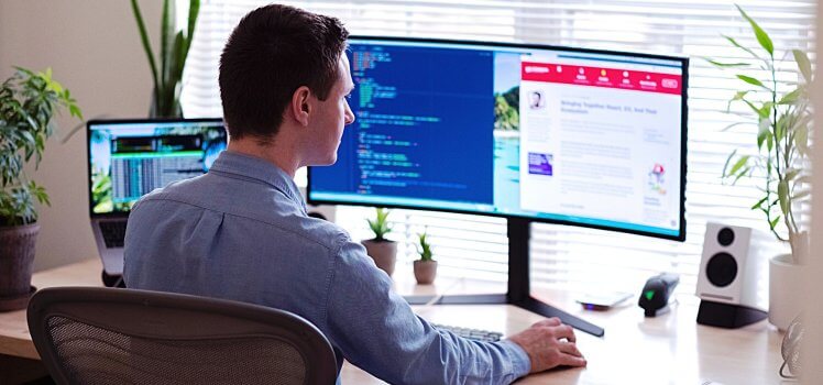 Man in a blue shirt working at a desk in front of a window with two large computer screens.