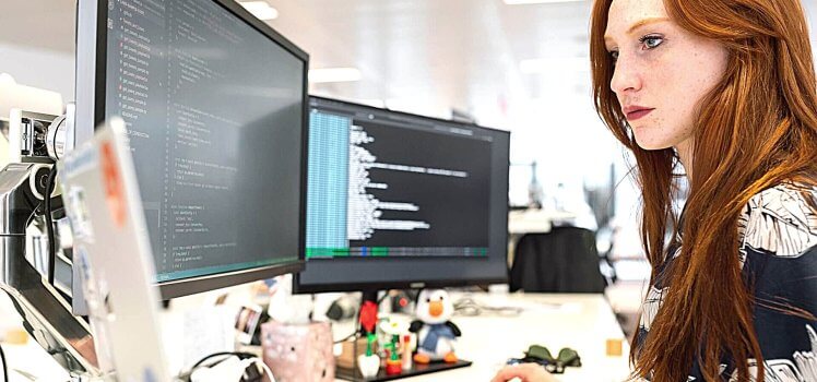 Side profile of a woman with red hair working at a desk with three large screens.