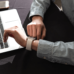 Person in a jean shirt looking at their white watch while sitting in front of a computer and cell phone.