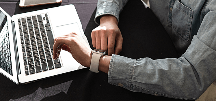 Person in a jean shirt looking at their white watch while sitting in front of a computer and cell phone.