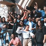 Large group of people with hands in the air posing for a picture in front of a stone building.