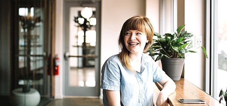 Woman in a blue shirt sitting at a tall bar in front of a window smiling.