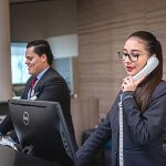 Two individuals working behind a hotel desk. One woman holding a phone at a computer.