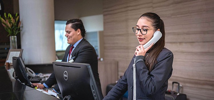 Two individuals working behind a hotel desk. One woman holding a phone at a computer.