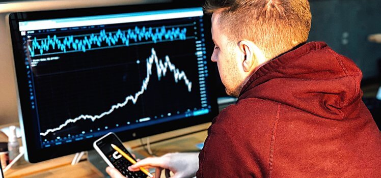 Man in a red hoodie looking at a phone screen in front of a large desktop screen.