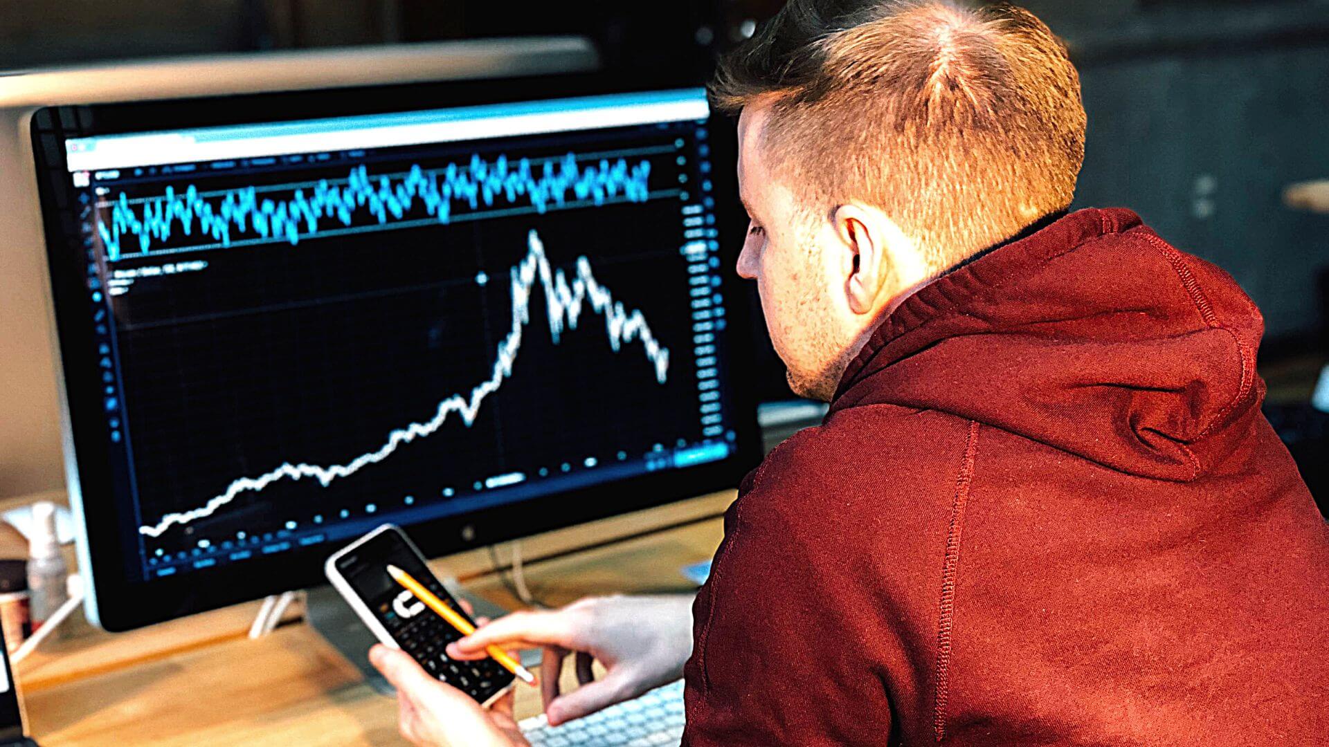 Man in a red hoodie looking at a phone screen in front of a large desktop screen.