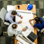 Overhead view of three people marking a building schematic displayed on a large table.