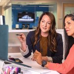 Two women sitting at one desk reviewing something on a blue laptop.