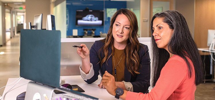 Two women sitting at one desk reviewing something on a blue laptop.