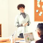 Woman with curly hair and glasses presenting to a group of three others in front of a peg board with sticky notes.