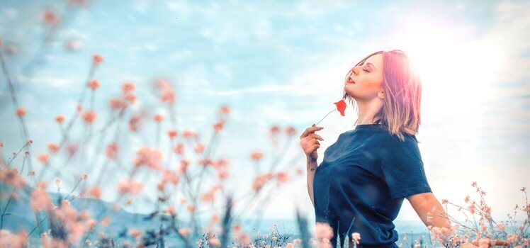 Woman in a blue shirt standing in a field of pink flowers holding a red flower facing the sky.