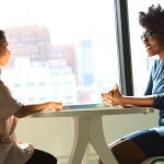 Two women sitting at a small table in front of windows discussing how to Improve mental health in the Workplace.