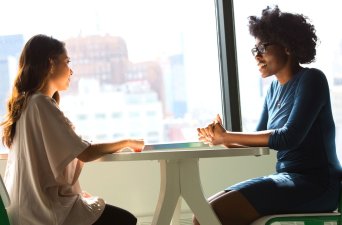 Two women sitting at a small table in front of windows discussing how to Improve mental health in the Workplace.