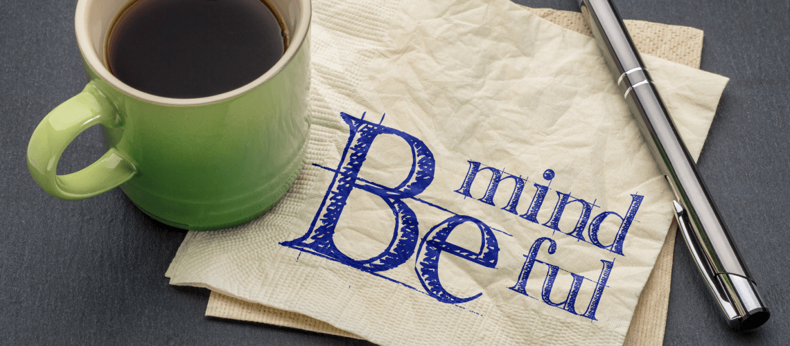 Coffee cup and pen sitting on a table with a napkin that reads Be Mindful.
