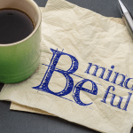 Coffee cup and pen sitting on a table with a napkin that reads Be Mindful.