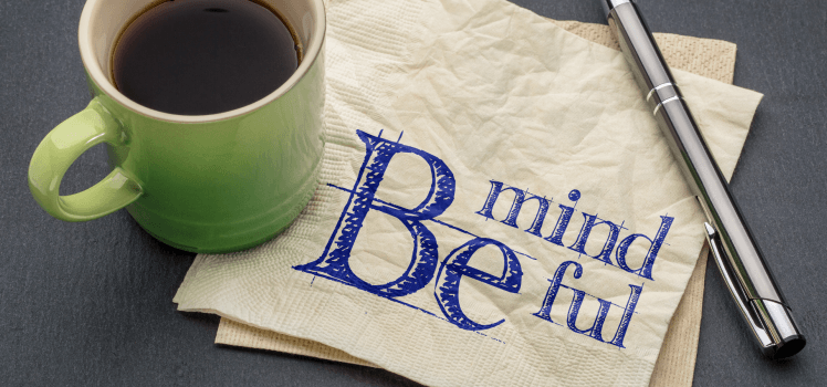 Coffee cup and pen sitting on a table with a napkin that reads Be Mindful.