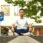 Man in a white shirt sitting on a concrete block meditating on a city street.