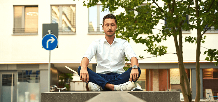 Man in a white shirt sitting on a concrete block meditating on a city street.