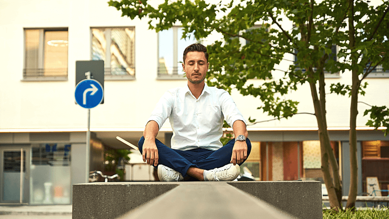 Man in a white shirt sitting on a concrete block meditating on a city street.