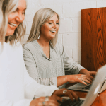 Two women sitting at a small table working on laptops.