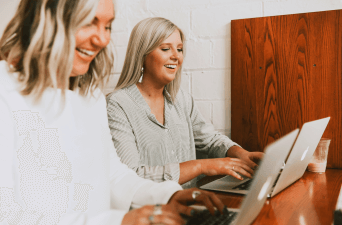 Two women sitting at a small table working on laptops.