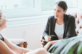 Two women sitting closely having a conversation in a sunlight filled room.