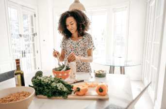 Woman in a bright dining room making a salad in an orange salad bowl.