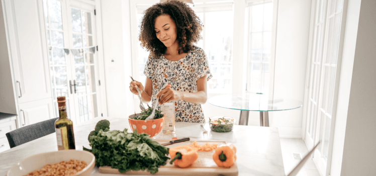 Woman in a bright dining room making a salad in an orange salad bowl.