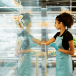 Woman in a blue apron restocking a clear grocery store display.