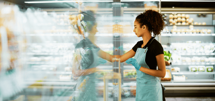 Woman in a blue apron restocking a clear grocery store display.