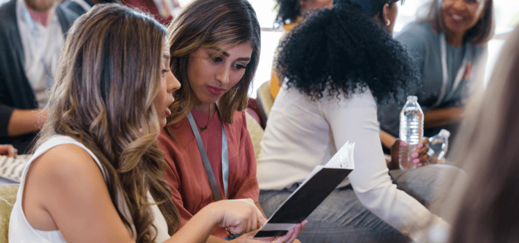 Two women conversing and looking at a pamphlet in a large group.