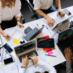 Overhead perspective of a table full of workers on laptops and writing on paper.