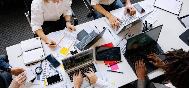 Overhead perspective of a table full of workers on laptops and writing on paper.