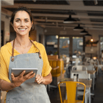 Woman in a yellow shirt standing in front of a cafe working on a tablet smiling.