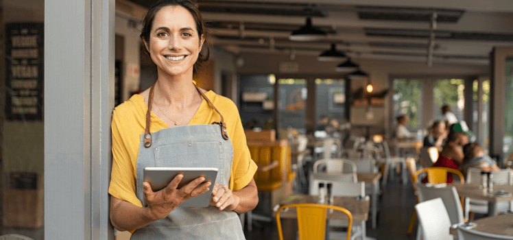 Woman in a yellow shirt standing in front of a cafe working on a tablet smiling.