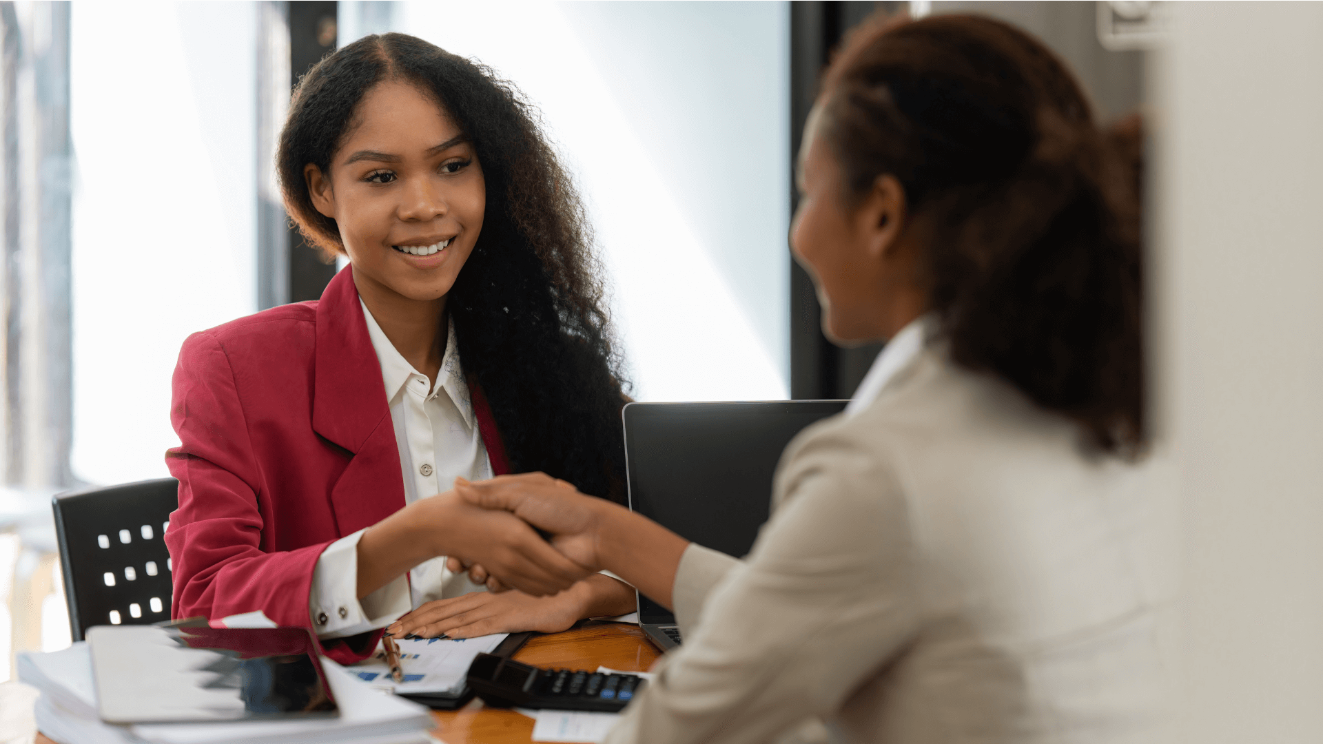 Two women shaking hands over a table in an interview setting.