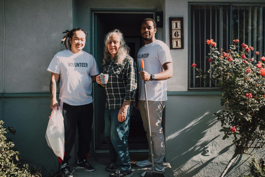 Two men assisting and older woman with sweeping and picking up trash while they volunteer.